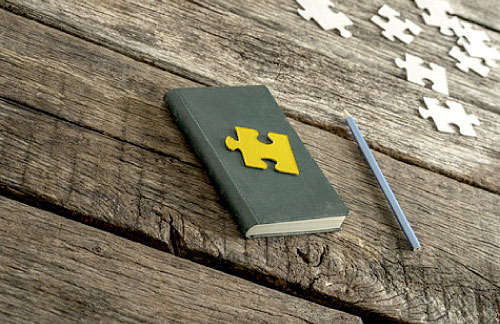 Viele weisse Puzzleteile. Auf einem Notizbuch liegt ein gelbes Puzzleteil.